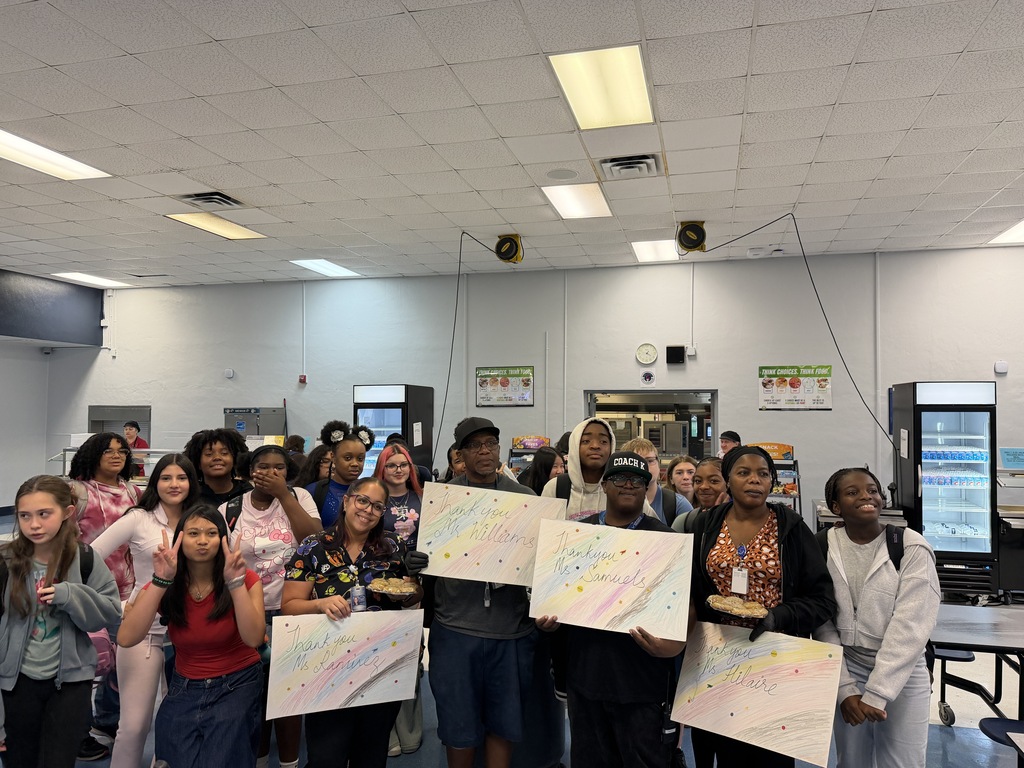 A group of people in a classroom, holding up multicolored papers with drawings. A refrigerator is visible in the background.