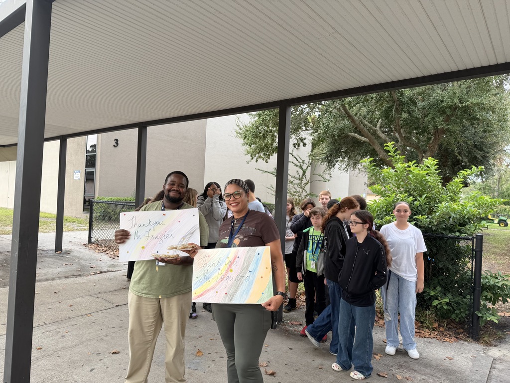People gather in a shaded area, holding colorful posters. Two individuals stand out, one holding a large poster. Background features a building and greenery.