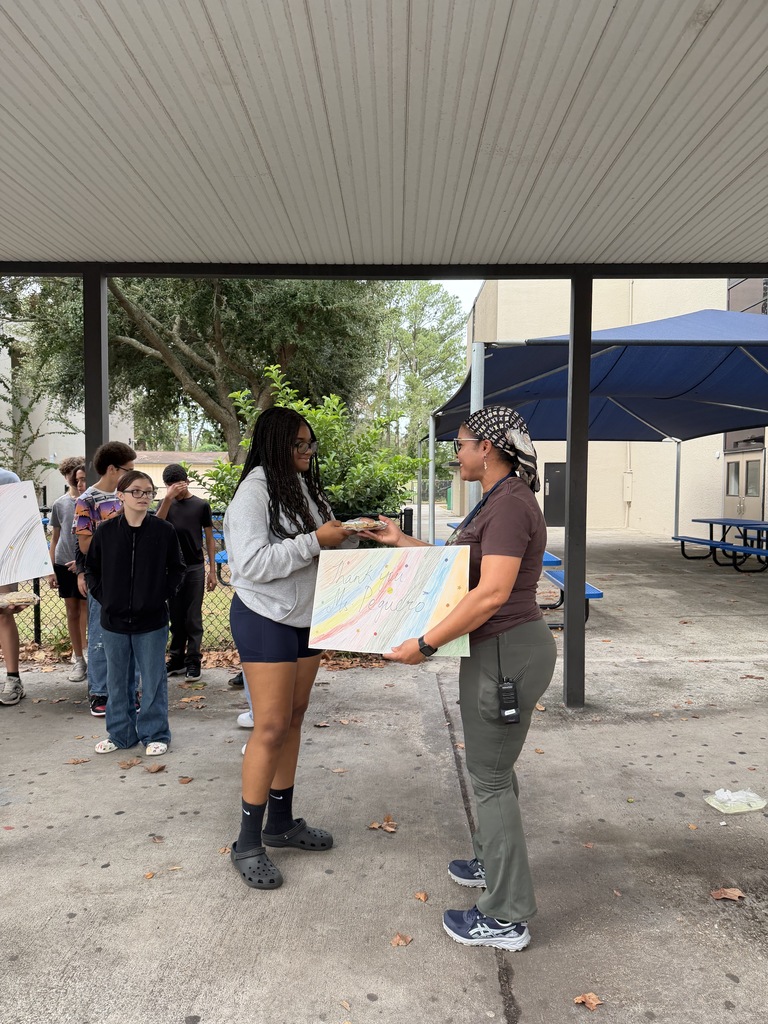 Two women stand under an awning. One holds a map and hands it to the other. People stand behind them. Trees and buildings are in the background.