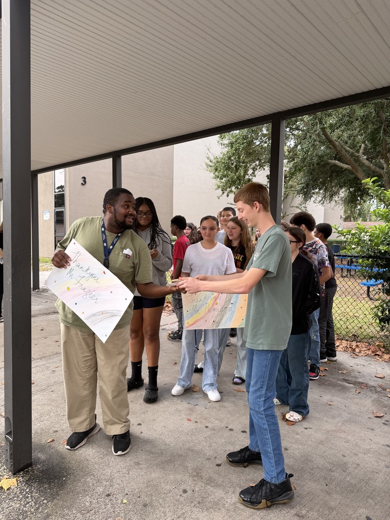 Group of people stand outside with two people holding papers. One is handing a paper to another.