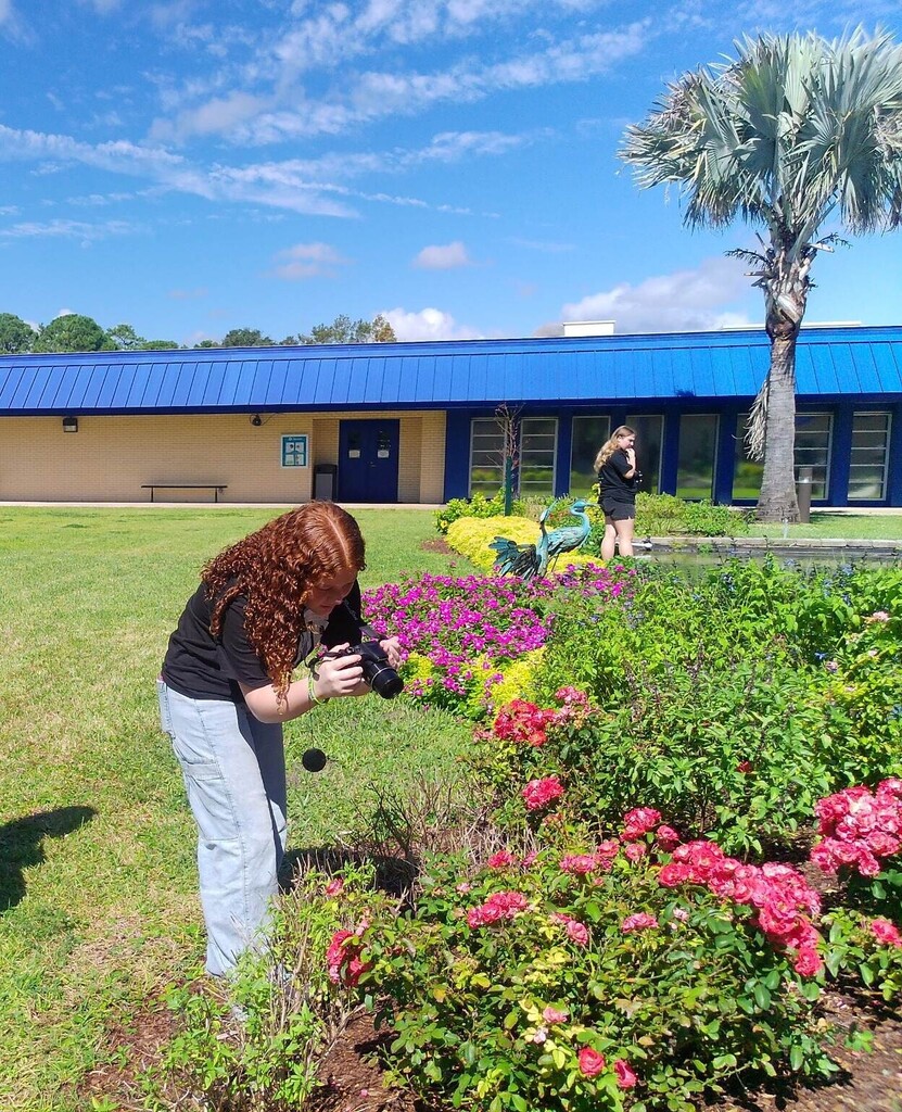 A person takes photos of flowers in a garden. Behind them, a blue building is visible.