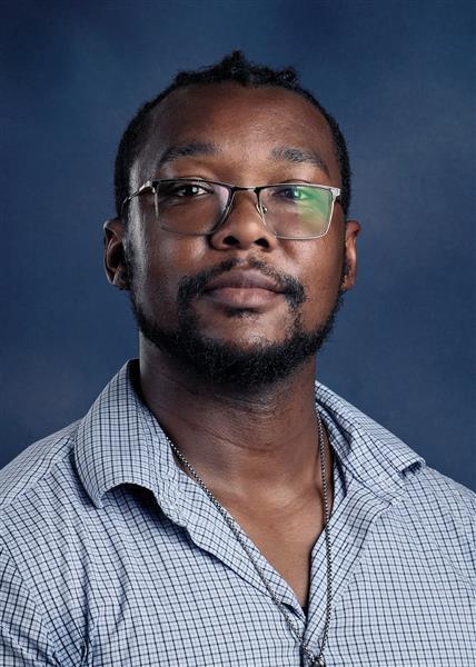 A man with a beard and glasses stands against a blue background. He wears a blue-and-white checkered shirt.