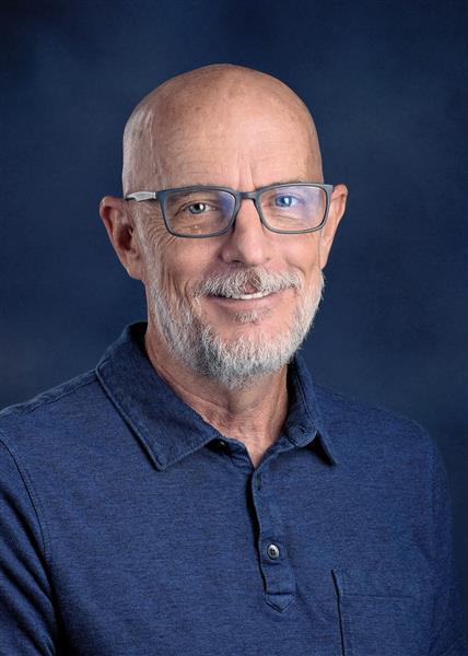 A man with a white beard, wearing glasses and a blue polo shirt, smiles against a blue background.