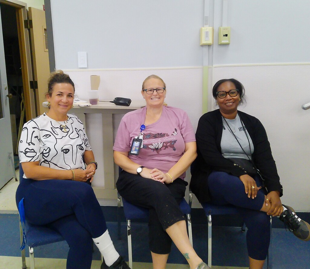 Three women sit on blue chairs in a room. Two wear glasses. One wears a purple shirt with a name tag.