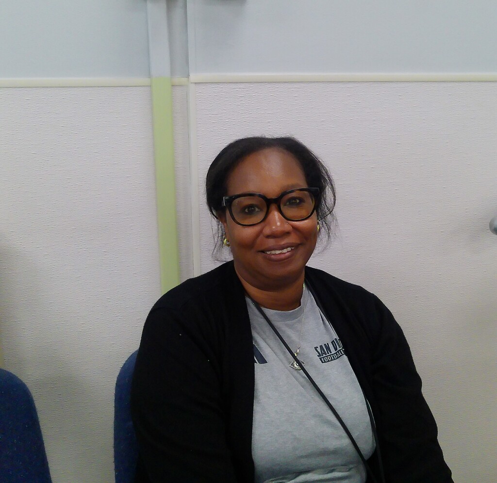 A woman sits in a blue chair against a white wall, smiling. She wears glasses, a gray shirt, and a black cardigan.