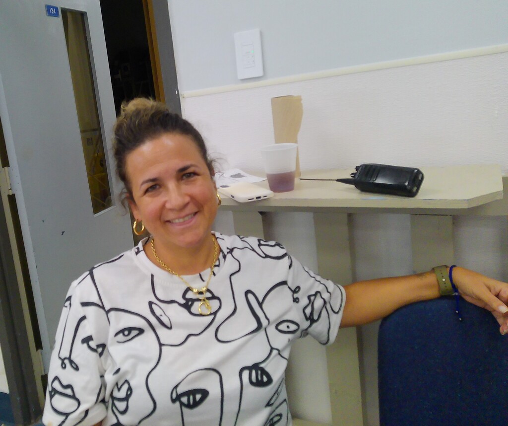 Woman smiling, seated in an office, wearing a white shirt with abstract black images. Behind her is a white desk with a cup and a black object.