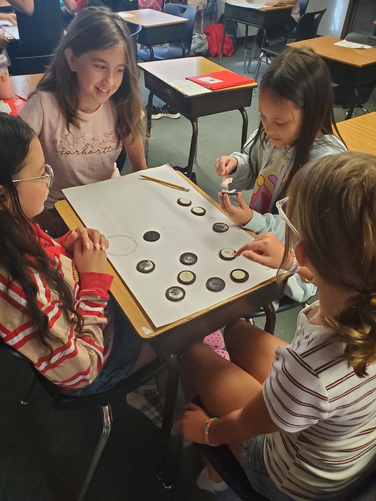 Four girls sit at a table, working with a board game and playing pieces in a classroom.