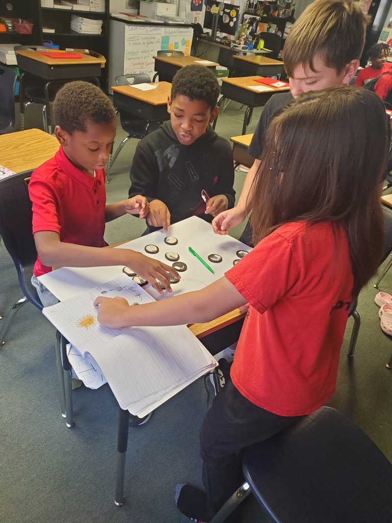 Four children at a table, focusing on an activity. One child holds a marker. A white paper with colored markers and a ruler is on the table.