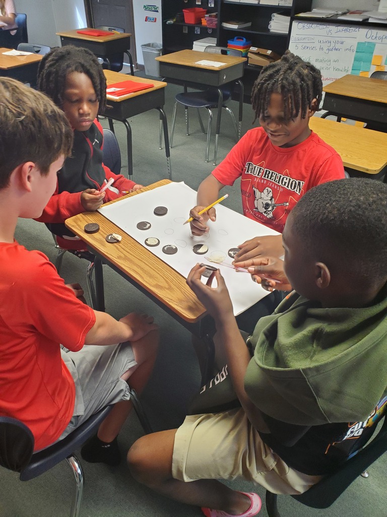 Four children sit around a desk, each holding a marker and working on a paper.