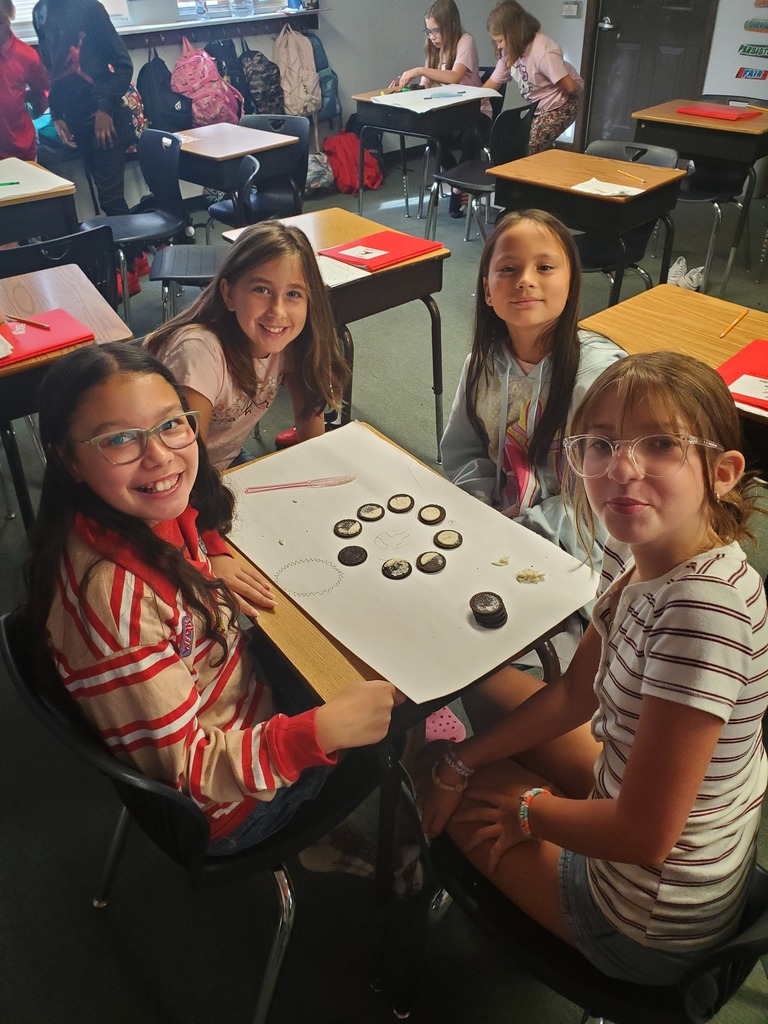 Four girls are sitting at a desk in a classroom, smiling and interacting with items on a table.