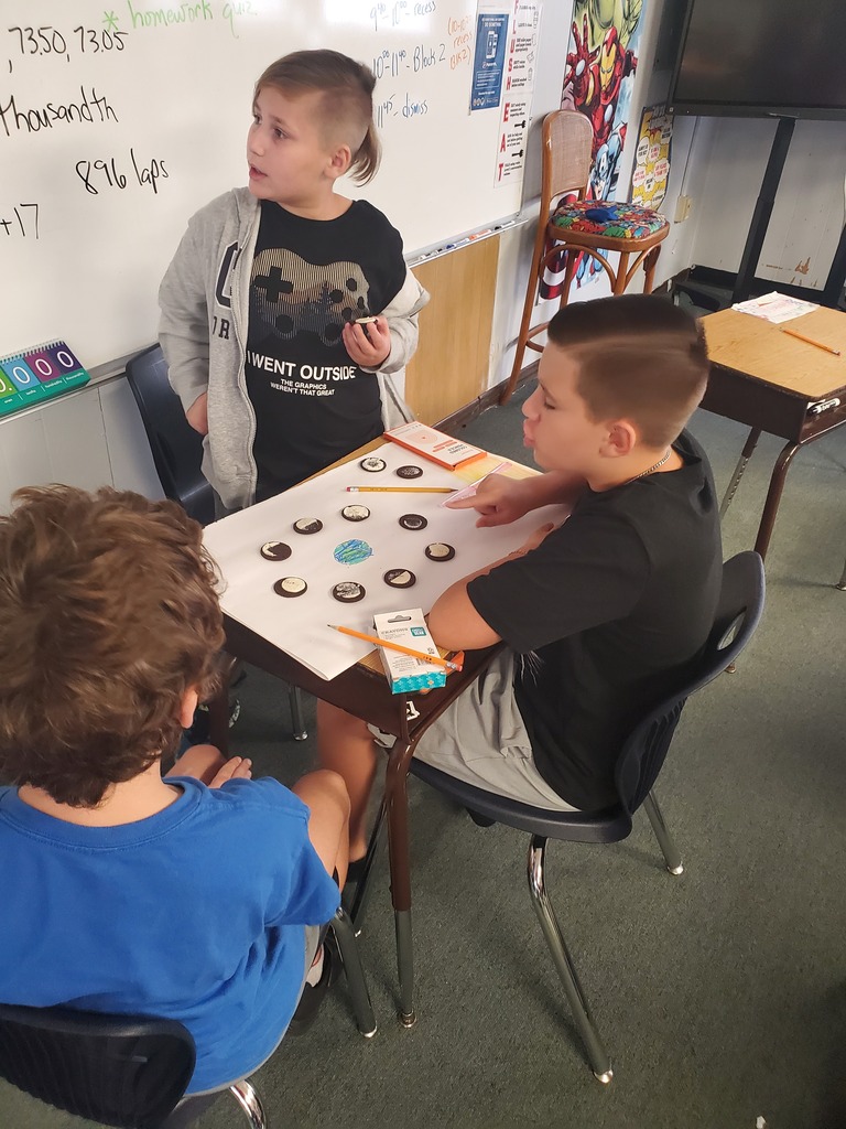 Three children seated at a table in a classroom. The boy in the middle is looking at a board with colored circles.