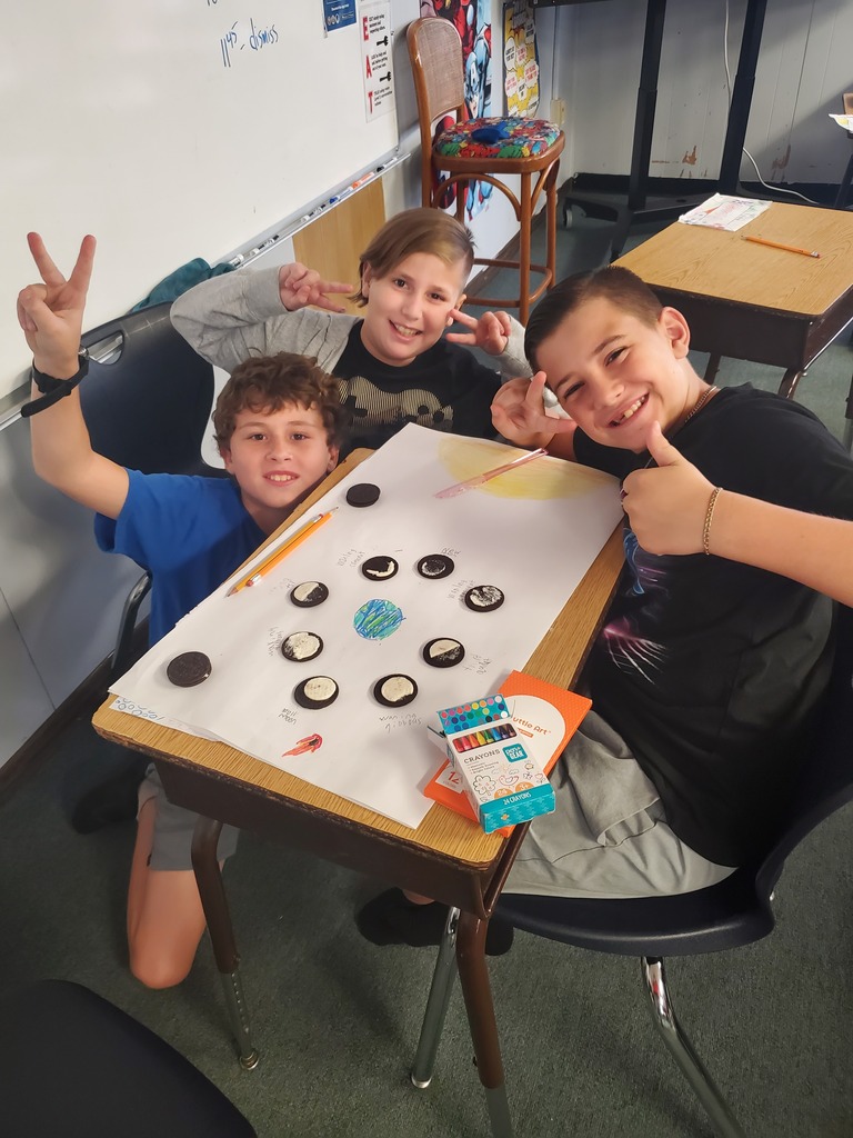 Three boys seated at a desk, smiling and making gestures. A table with cookies on it.