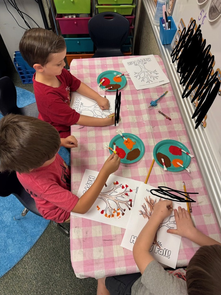 Three children are seated at a table, working on art projects. Each child holds a marker.