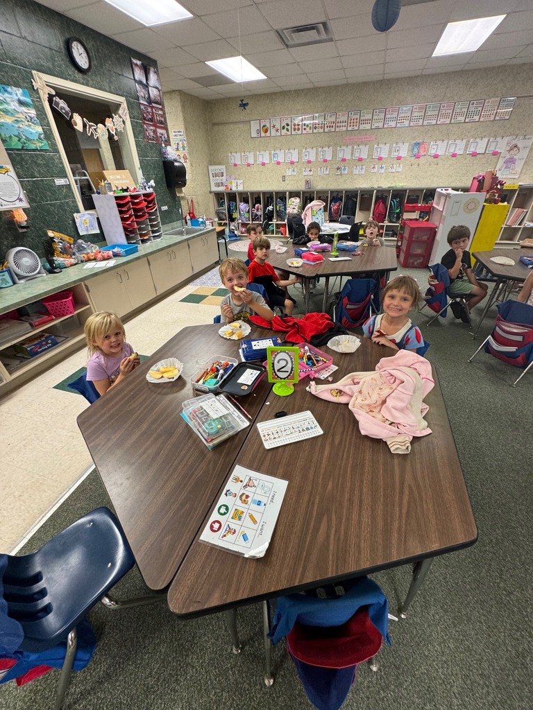 Small children seated at a table with books, crayons, and paper plates. Background has shelves and tables.