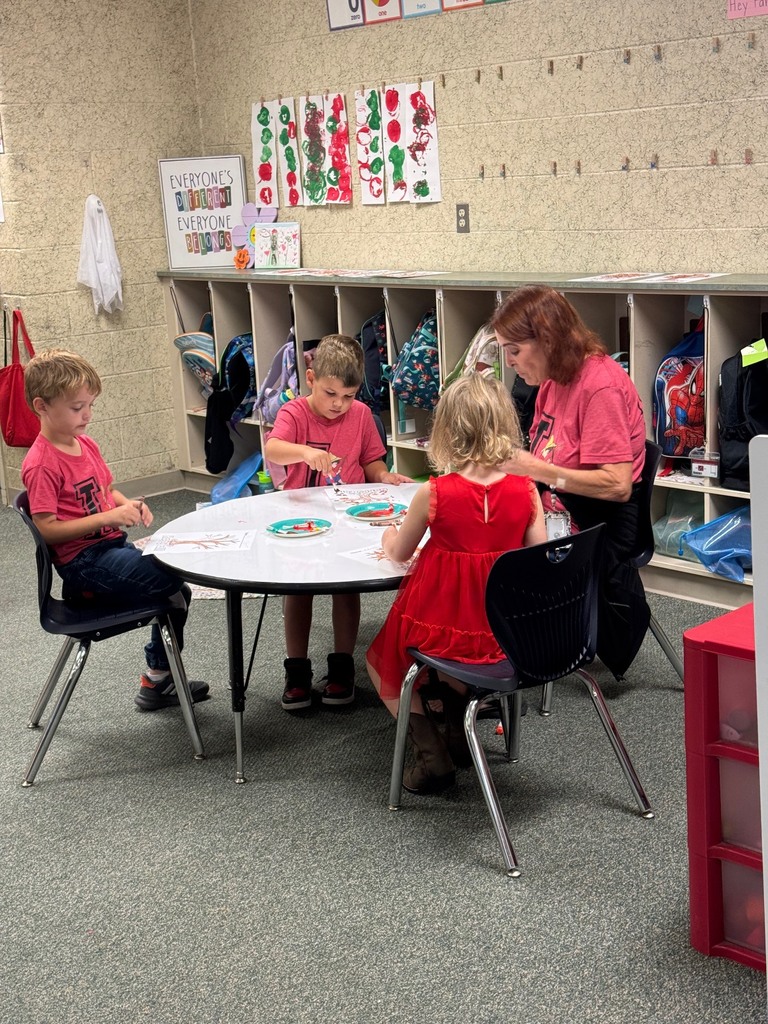 Four children in various clothes sit at a round table with an adult. Behind them are cubbies with bags and shirts.