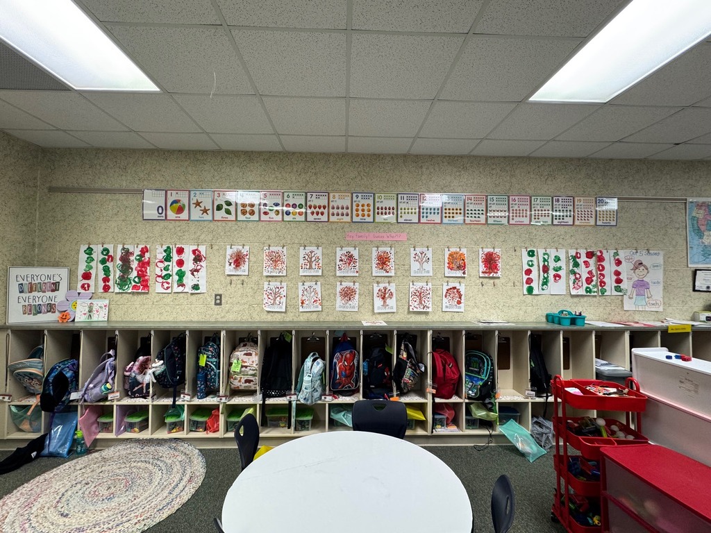 Interior of a classroom with light beige walls. A round table and chairs are in the foreground. Storage cubbies hold backpacks and other items.