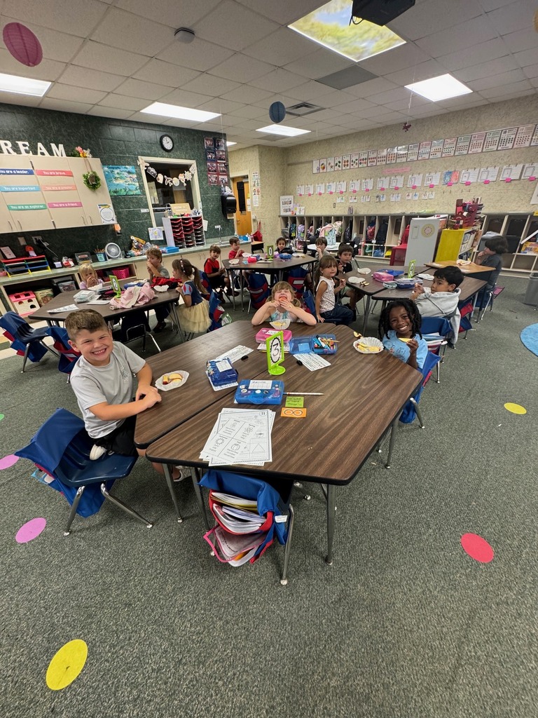 Children are seated at desks in a classroom. Some are focused on tasks, while others look up. The room has tables and chairs, and colorful dots decorate the floor.