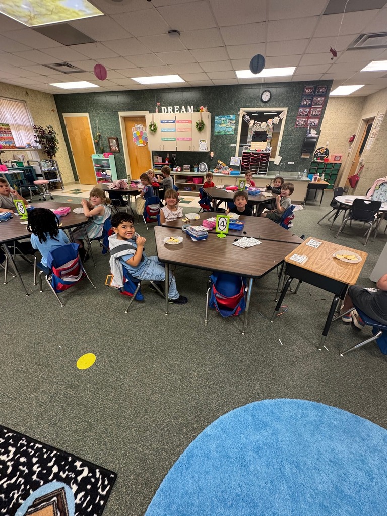 Classroom with students seated at tables. Tables are set with plates, cups, and utensils. Bright overhead lighting.