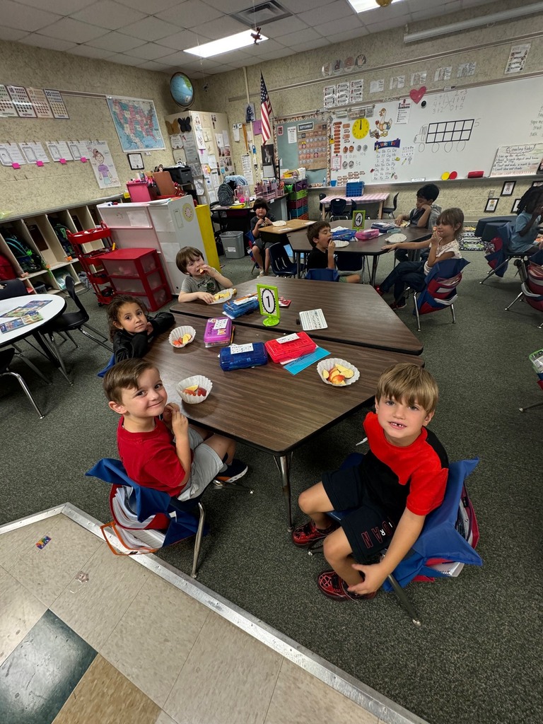 Children seated at tables in a classroom. One child has a book, and another has a plate.