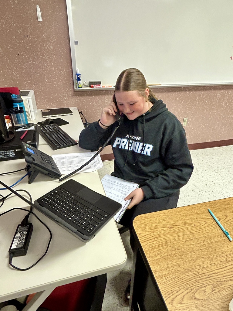 Person sitting at desk, holding a phone, wearing a black hoodie with "Premier" text, surrounded by papers and a laptop.