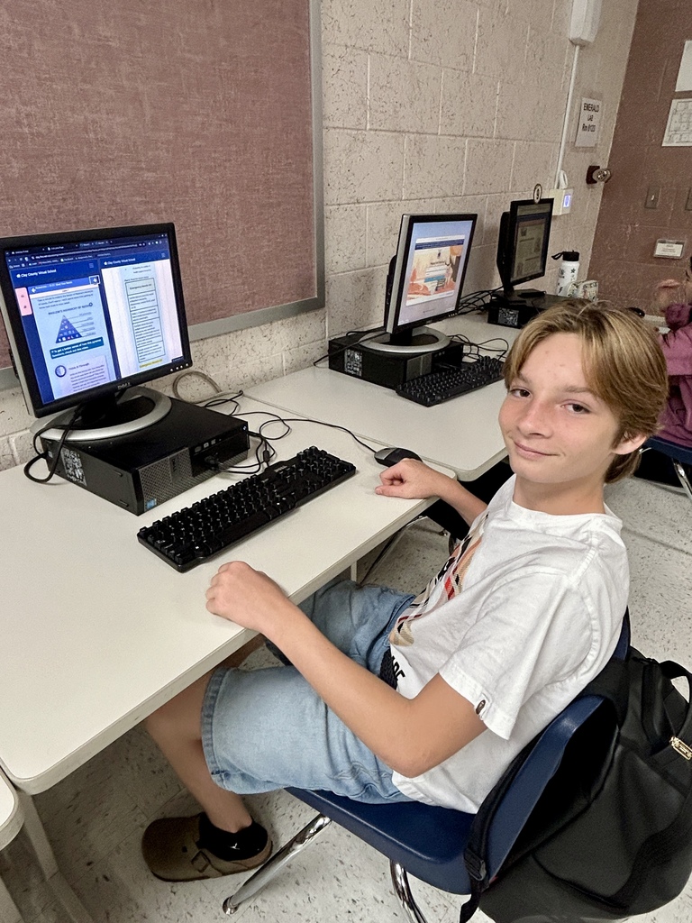 A young person sits at a desk, smiling, with two computer monitors, a keyboard, and a mouse.