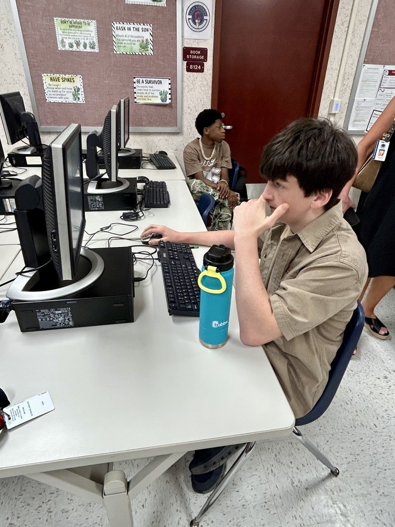 A person seated at a desk, using a computer, with a blue tumbler nearby. Another person sits in the background.