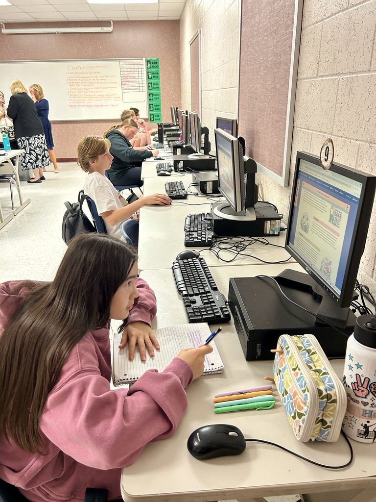 Students seated at computers in a classroom, one with a mouse and a notebook, and a water bottle nearby.
