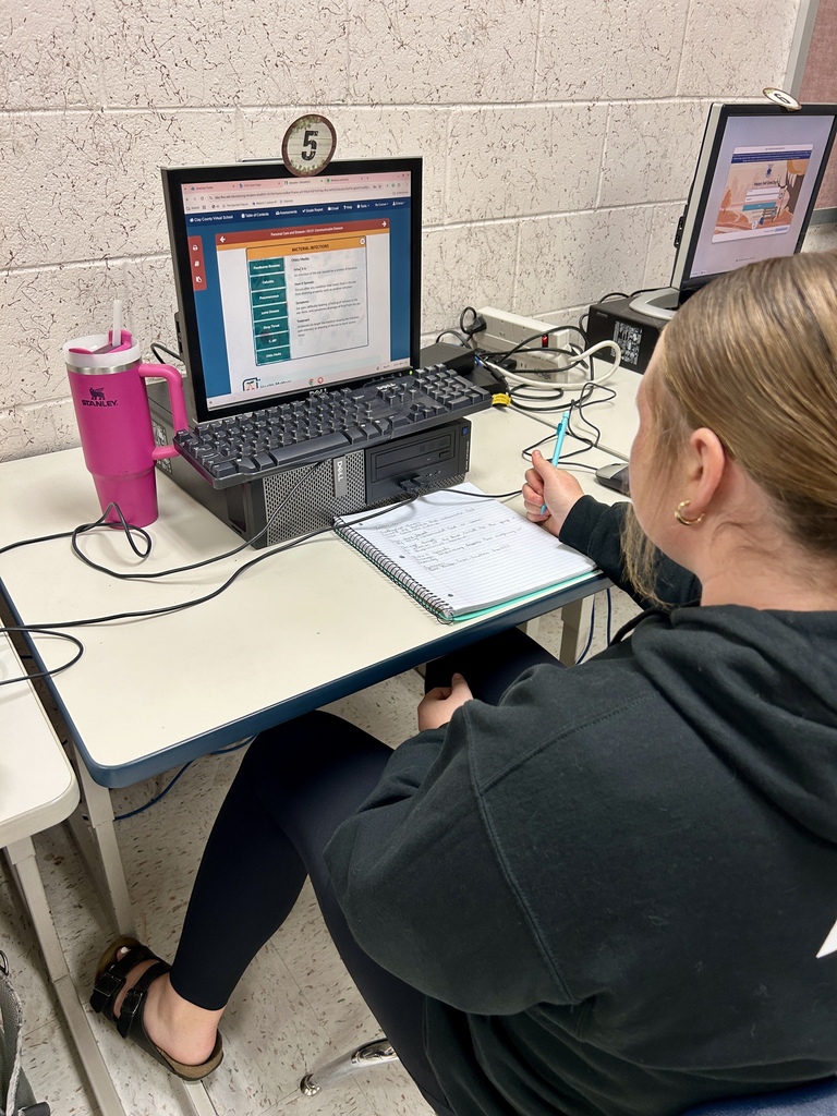 A person sits at a desk with a laptop, pen, and notebook. A pink tumbler rests on the desk.
