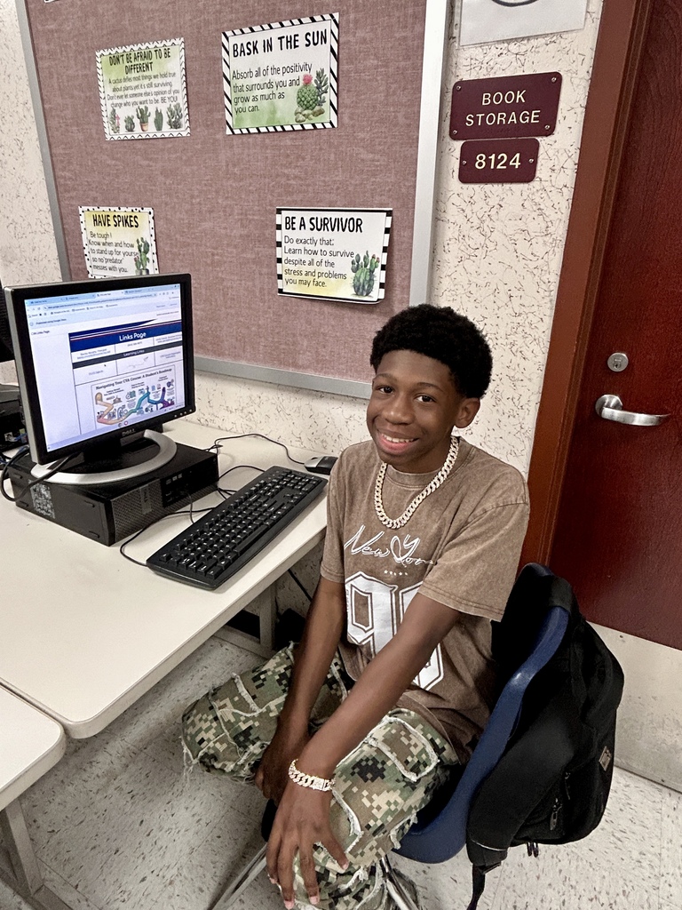 A boy with a necklace sits in a chair at a desk with a computer.