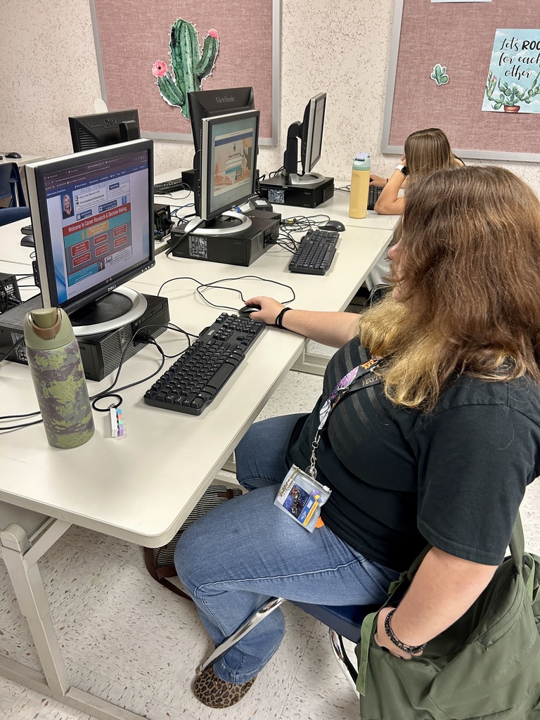 A woman sits at a desk, using a computer while wearing a black shirt, blue jeans, and sneakers.