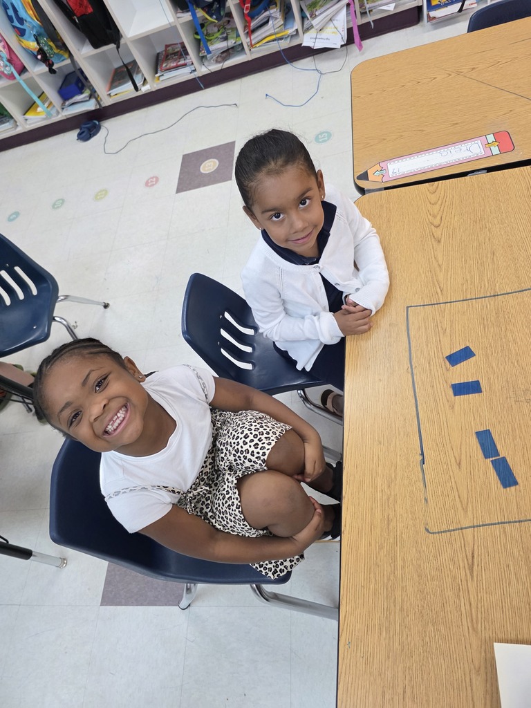 Two children in white shirts sit at a table in a classroom with blue chairs and a white floor.