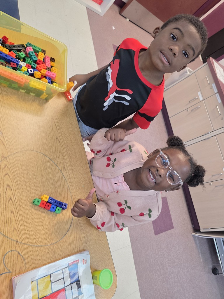 Two children standing at a table, one with glasses, both smiling. A yellow box with multicolored blocks on the table.