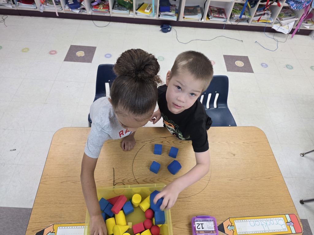 Two students build numbers with colorful cubes on a wooden table in a classroom.