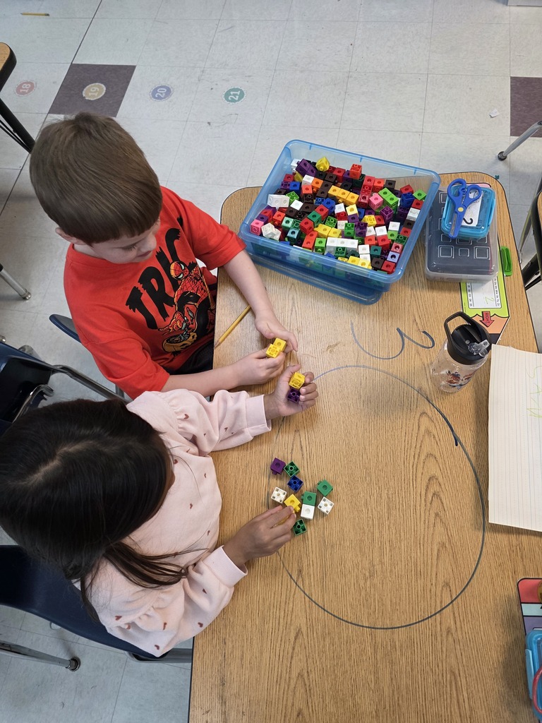 Two children in a classroom, seated at a table, engaged with multi-colored cubes, possibly involved in a learning activity.