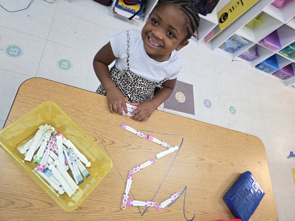 A child in a classroom smiles and plays with a yellow container of markers on a table.