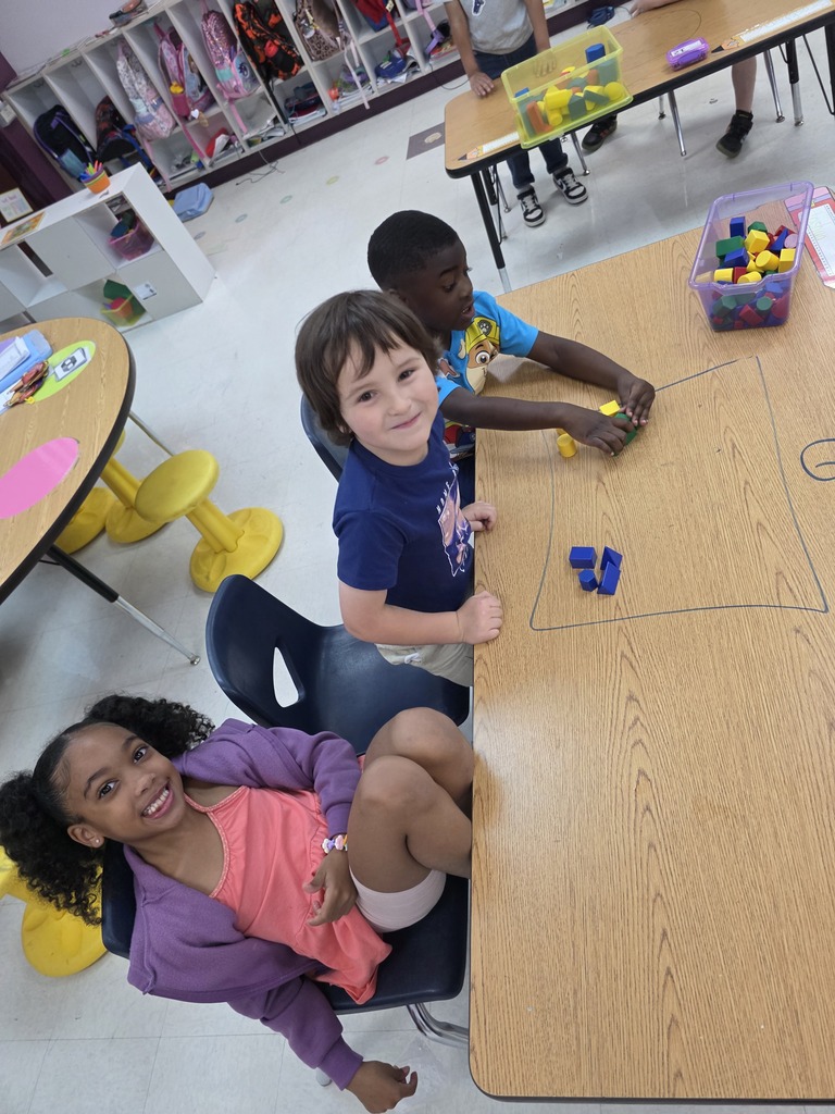 Three children are seated around a table, engaged with colorful building blocks in a classroom setting.