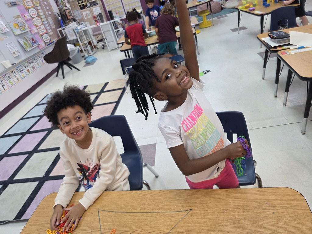 Two children sit at a table with colorful shapes. Behind them, several students are seated at tables. The room has white walls and a white floor.