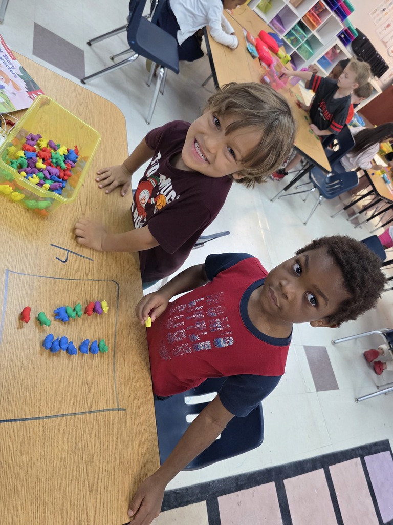 Two children, one with a red shirt and the other with a maroon shirt, sit at a table, smiling and playing with colorful blocks.