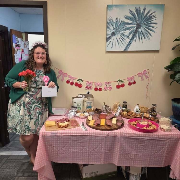 A woman stands by a table with a pink tablecloth, holding a certificate. The table displays a variety of foods.