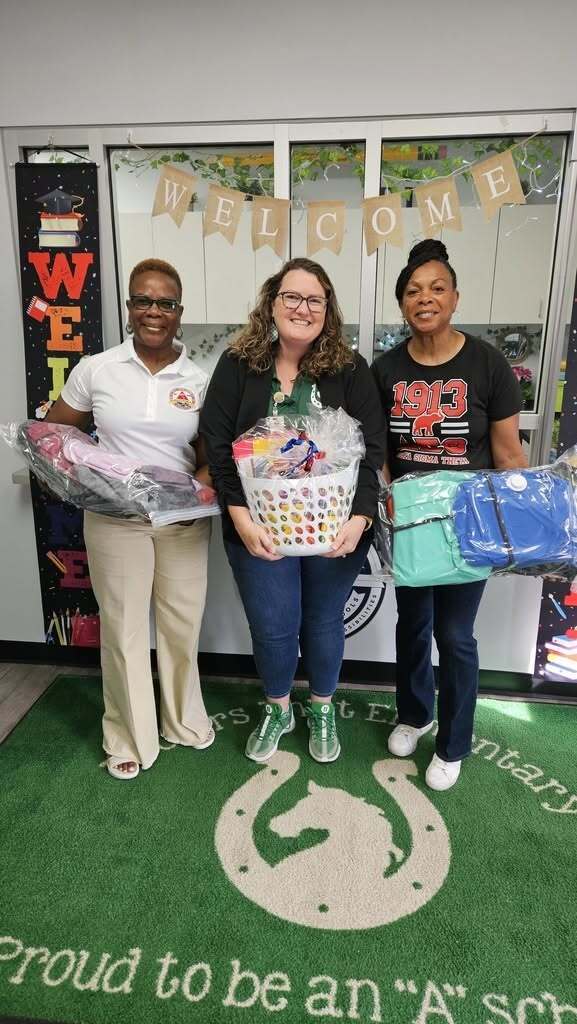 Three women pose in front of a welcome sign holding baskets. The woman on the left holds wrapped items.
