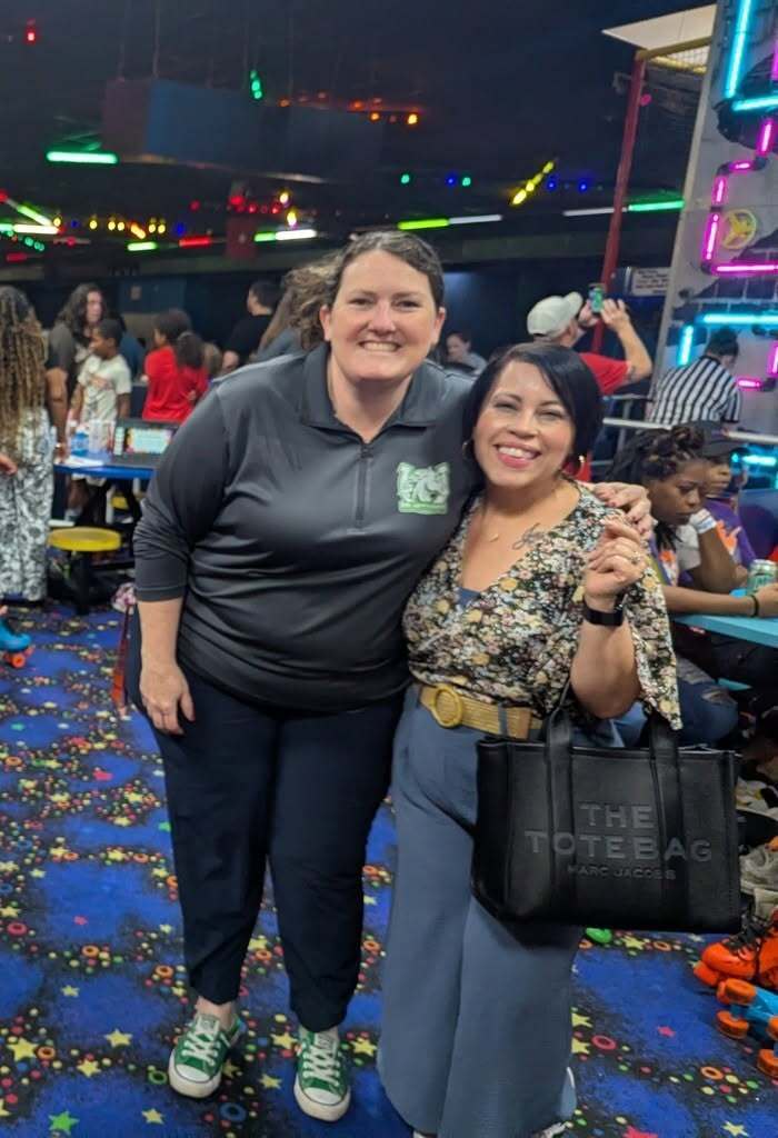 Two women stand in a brightly lit room, smiling and posing for a photo, with a colorful floor and decorative lights.