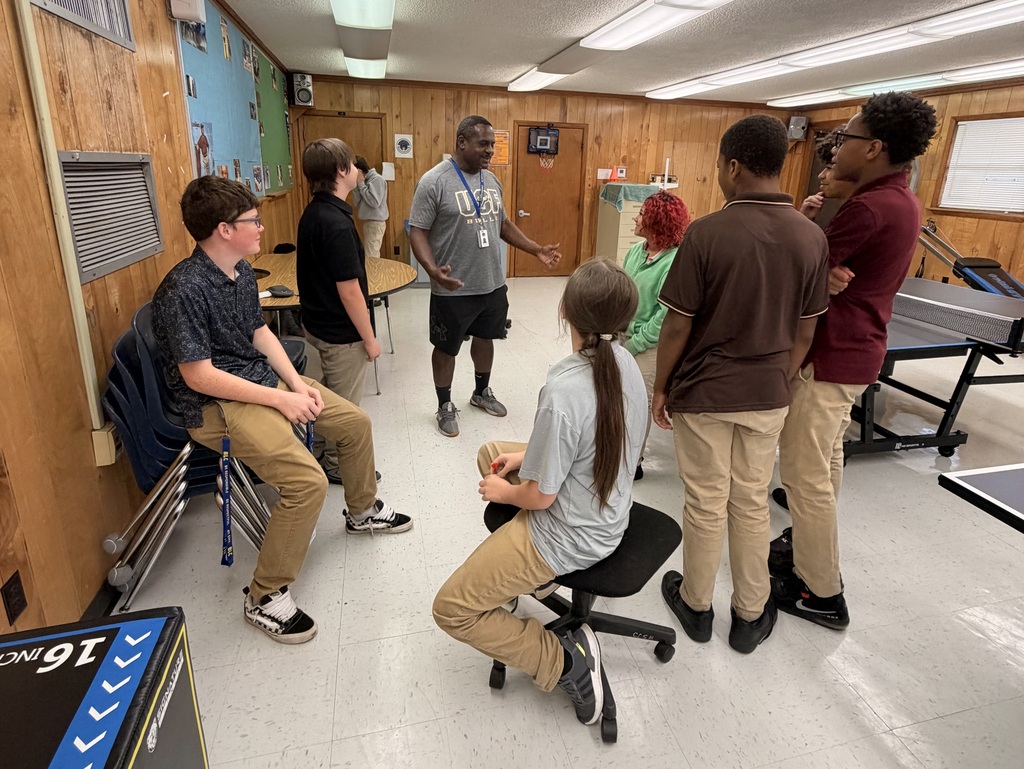 Several young people in a room. Some stand and others sit. One person is talking. A ping pong table is visible.