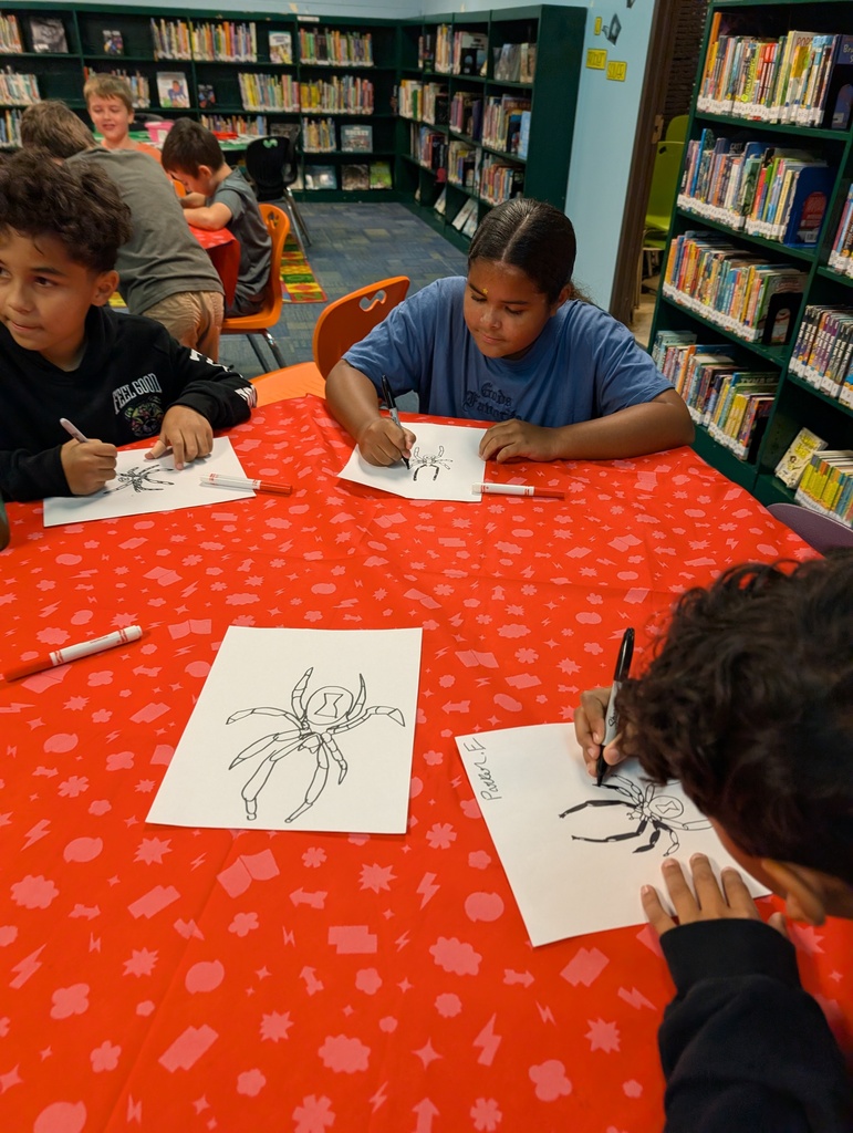 Four children draw at a table covered with a red tablecloth with spider images. They sit in a room with bookshelves and other people.