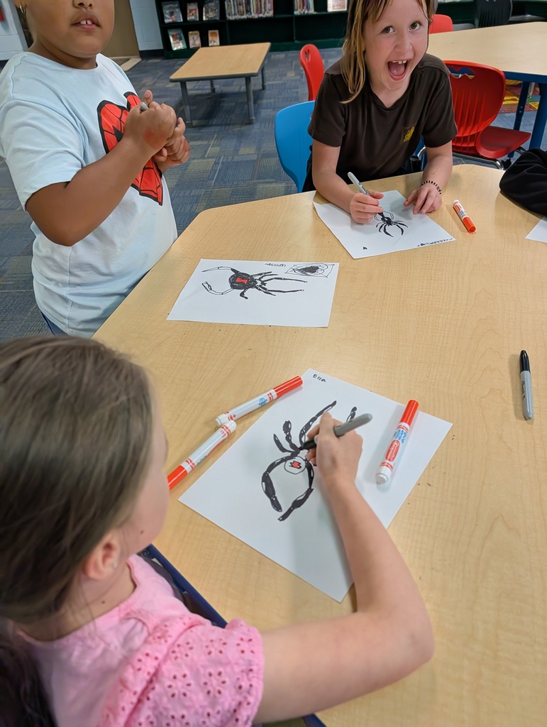 Children seated at a table, one drawing a spider on paper, the other holding a marker. Background includes chairs and tables.