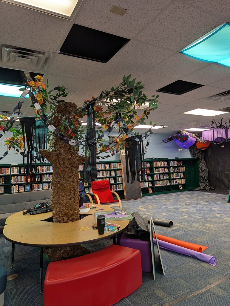 A library with bookshelves. An artificial tree is at the center, surrounded by a desk with a mug and a red chair.
