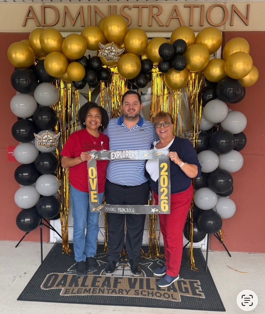 Three people stand in front of a balloon arch, holding signs that read "OVE" and "2026." They are in front of an Oakleaf Village Elementary School sign.