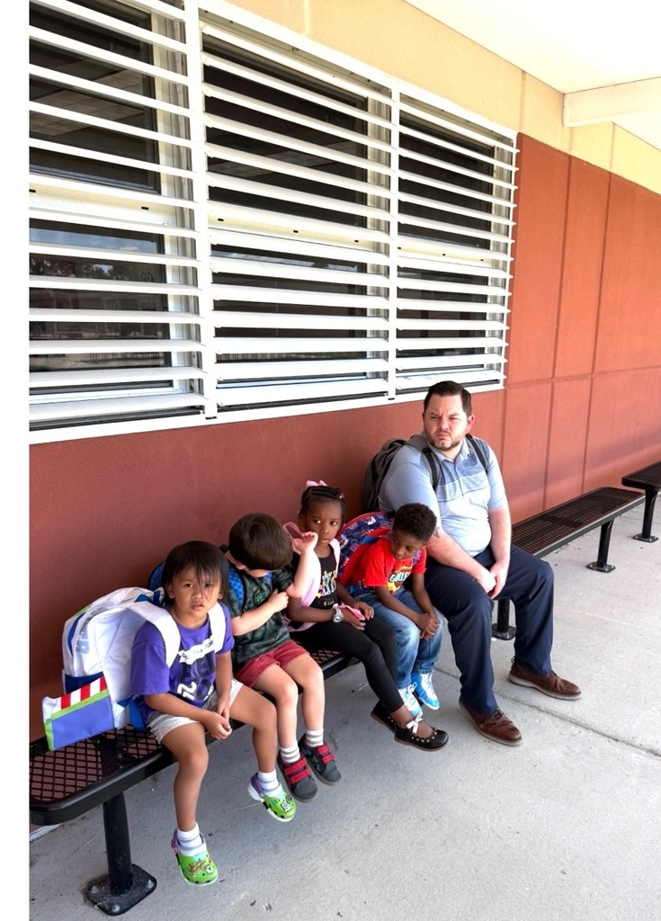 Group of children and Mr. Martin sit on a bench outside a building with closed windows.
