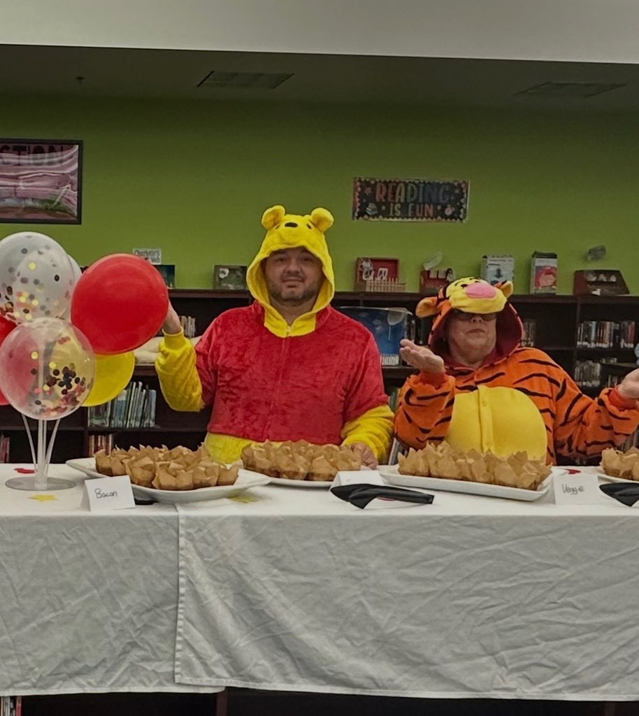 Two people in costumes sit at a table with food and balloons in front of a bookshelf.