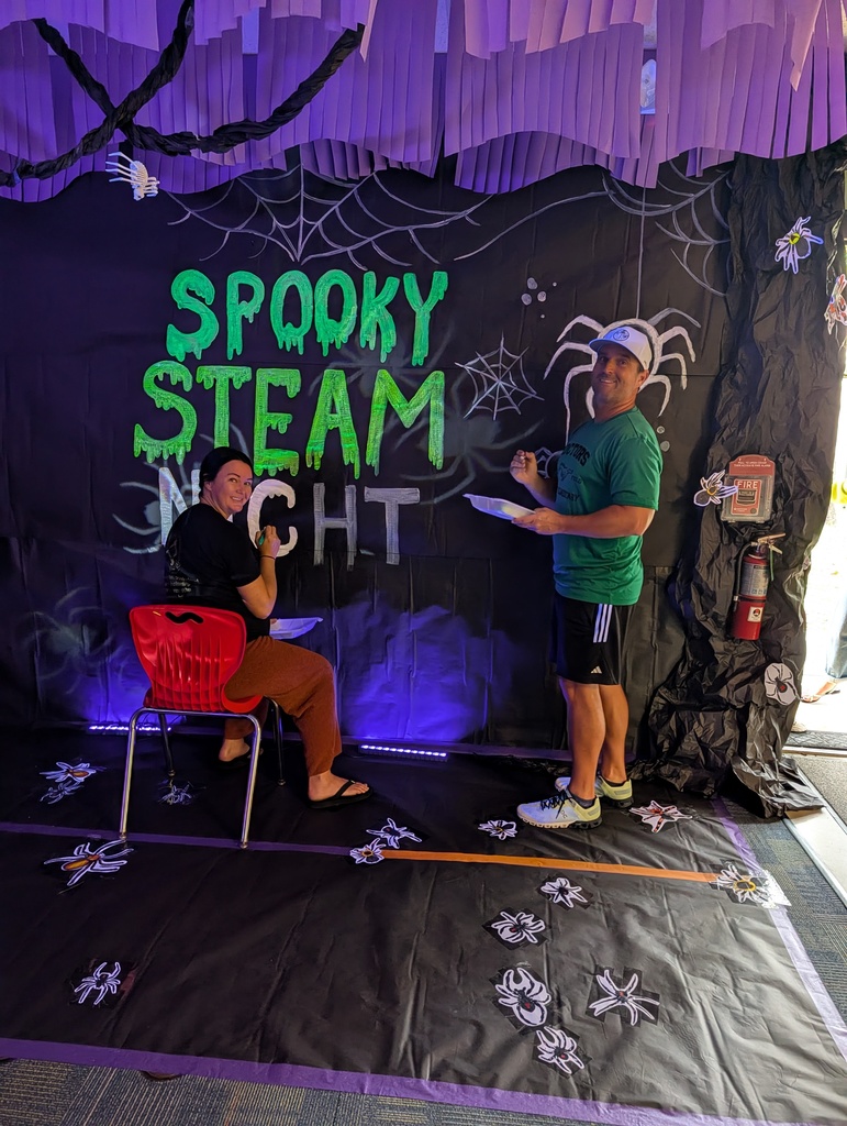 Two people are on a stage, one writing on a whiteboard. They are in front of a backdrop that says "Spooky Steam Night".