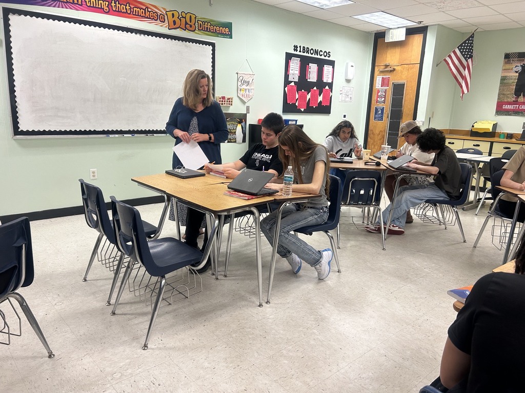 A classroom with a teacher standing next to a whiteboard. Students are seated at desks, using laptops.